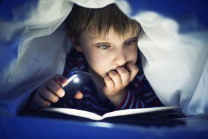 Little boy aged 5 is secretly reading book under sheets using mobile light. The book is very interesting and the boy is quite lost in it.
